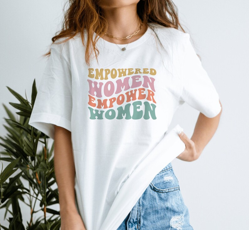 May include: White t-shirt with a graphic print that says "EMPOWERED WOMEN EMPOWER WOMEN" in a retro font with pink, yellow, and blue colors.