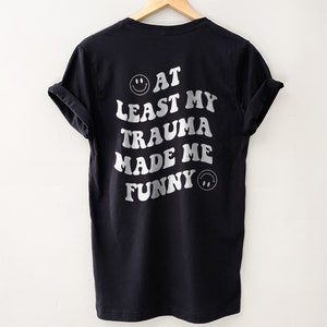 Op de afbeelding: Zwart T-shirt met witte tekst die "Tenminste mijn trauma heeft me grappig gemaakt" luidt, met een smiley aan beide kanten van de tekst.