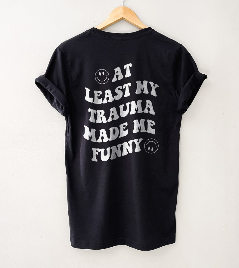 Op de afbeelding: Zwart T-shirt met witte tekst die "Tenminste mijn trauma heeft me grappig gemaakt" luidt, met een smiley aan beide kanten van de tekst.