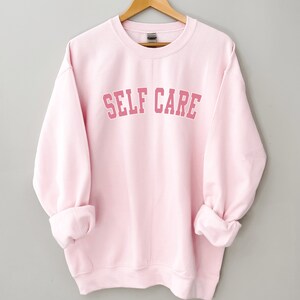 Peut inclure: Un sweat-shirt rose avec le texte "SELF CARE" dans une police d'&eacute;criture rose de style universitaire.