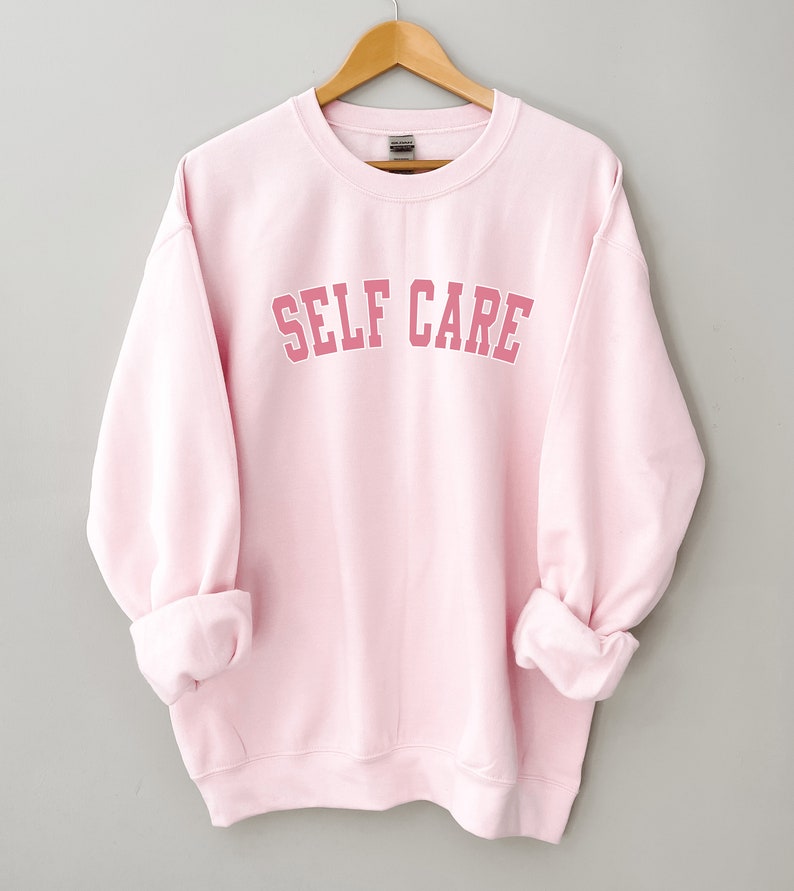 Peut inclure: Un sweat-shirt rose avec le texte "SELF CARE" dans une police d'&eacute;criture rose de style universitaire.