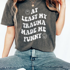 May include: A dark gray t-shirt with a white graphic that reads "At least my trauma made me funny" with a smiley face on either side of the text.
