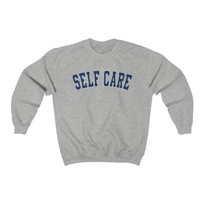 Peut inclure: Un sweat-shirt gris avec le texte "SELF CARE" en lettres bleues marine.