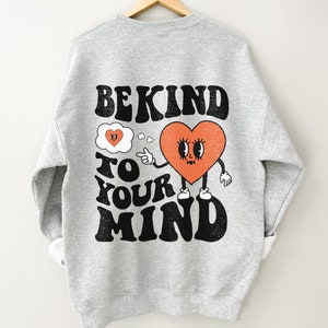 K&ouml;nnte beinhalten: Grauer Pullover mit einem schwarzen und orangefarbenen Grafikdruck, auf dem "Be Kind To Your Mind" steht. Die Grafik zeigt ein Herz mit einem Gesicht und eine Gedankenblase mit einem kleineren Herzen darin.