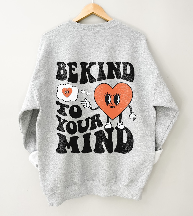 K&ouml;nnte beinhalten: Grauer Pullover mit einem schwarzen und orangefarbenen Grafikdruck, auf dem "Be Kind To Your Mind" steht. Die Grafik zeigt ein Herz mit einem Gesicht und eine Gedankenblase mit einem kleineren Herzen darin.