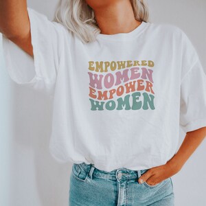 May include: White t-shirt with a graphic print that says "Empowered Women Empower Women" in a retro font with pink, yellow, and green colors.