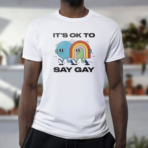 May include: White t-shirt with a graphic of two cartoon figures holding hands. The text "It's OK to Say Gay" is printed below the figures.