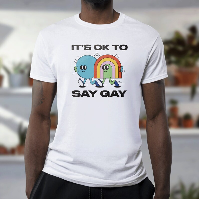 May include: White t-shirt with a graphic of two cartoon figures holding hands. The text "It's OK to Say Gay" is printed below the figures.