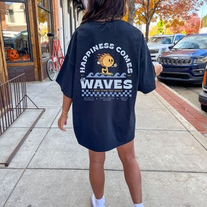 May include: Black t-shirt with the text "HAPPINESS COMES in WAVES" and a cartoon character surfing on waves. The shirt also includes the phrase "IT WILL FIND YOU AGAIN" and other text. The person is wearing white sneakers.