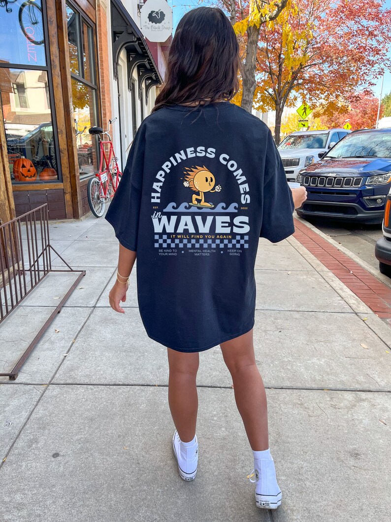 May include: Black t-shirt with the text "HAPPINESS COMES in WAVES" and a cartoon character surfing on waves. The shirt also includes the phrase "IT WILL FIND YOU AGAIN" and other text. The person is wearing white sneakers.