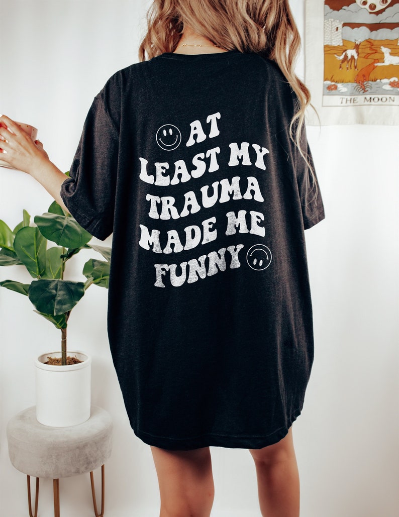 Op de afbeelding: Zwart oversized T-shirt met een witte grafische afbeelding met de tekst "At Least My Trauma Made Me Funny" met een smiley aan beide kanten van de tekst.
