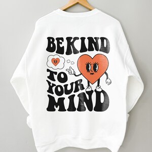 K&ouml;nnte beinhalten: Wei&szlig;er Pullover mit einem schwarzen grafischen Aufdruck, der "Be Kind To Your Mind" lautet. Die Grafik zeigt ein rotes Herz mit einem Gesicht und Armen, das auf eine Gedankenblase mit einem Herzen darin zeigt.
