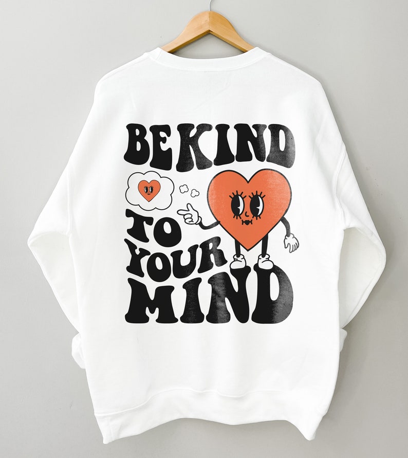 K&ouml;nnte beinhalten: Wei&szlig;er Pullover mit einem schwarzen grafischen Aufdruck, der "Be Kind To Your Mind" lautet. Die Grafik zeigt ein rotes Herz mit einem Gesicht und Armen, das auf eine Gedankenblase mit einem Herzen darin zeigt.