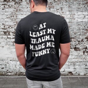 Op de afbeelding: Zwart T-shirt met witte tekst die "At Least My Trauma Made Me Funny" luidt met een smiley aan beide kanten van de tekst.