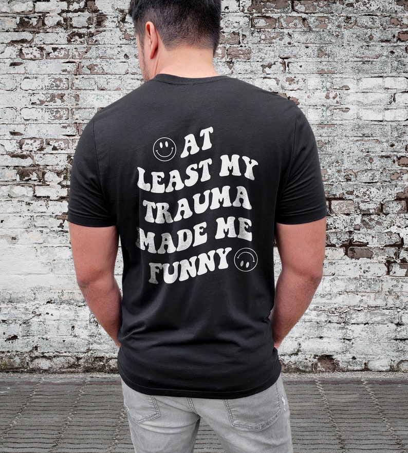 Op de afbeelding: Zwart T-shirt met witte tekst die "At Least My Trauma Made Me Funny" luidt met een smiley aan beide kanten van de tekst.