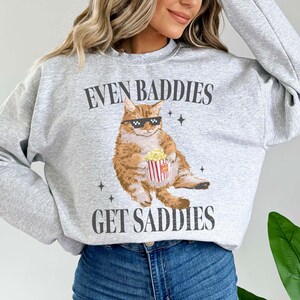 May include: Gray sweatshirt with a graphic of a cat wearing sunglasses and holding a bucket of popcorn. The text on the sweatshirt reads "Even Baddies Get Saddies".