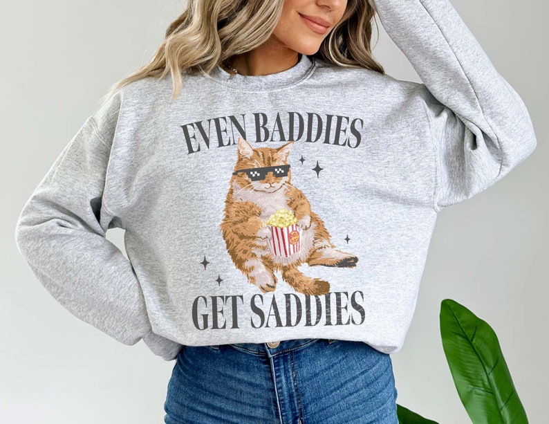 May include: Gray sweatshirt with a graphic of a cat wearing sunglasses and holding a bucket of popcorn. The text on the sweatshirt reads "Even Baddies Get Saddies".