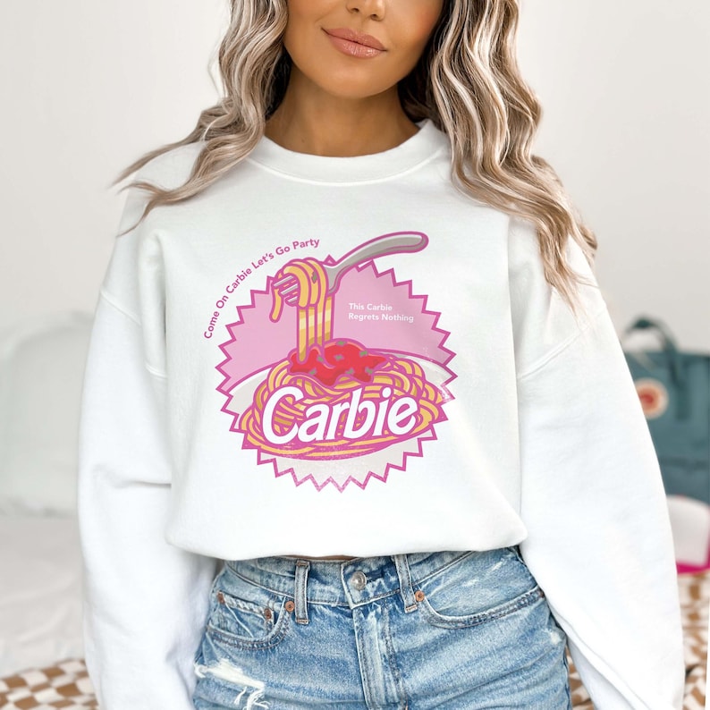 Może przedstawiać: Biała bluza z r&oacute;żowo-ż&oacute;łtym nadrukiem przedstawiającym talerz spaghetti z tekstem "Come On Carbie Let's Go Party" i "This Carbie Regrets Nothing" oraz słowem "Carbie" w dużym rozmiarze czcionki.