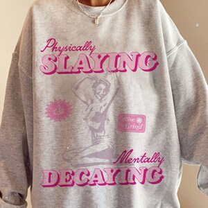 May include: Gray sweatshirt with a pink graphic print that says "Physically Slayin'" and "Mentally Decaying". The graphic also includes a silhouette of a woman in a dance pose and a small graphic that says "Rise & Grind".