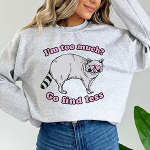 Pu&ograve; includere: Felpa grigia con un procione che indossa occhiali da sole a forma di cuore rosa e il testo "I'm too much? Go find less".
