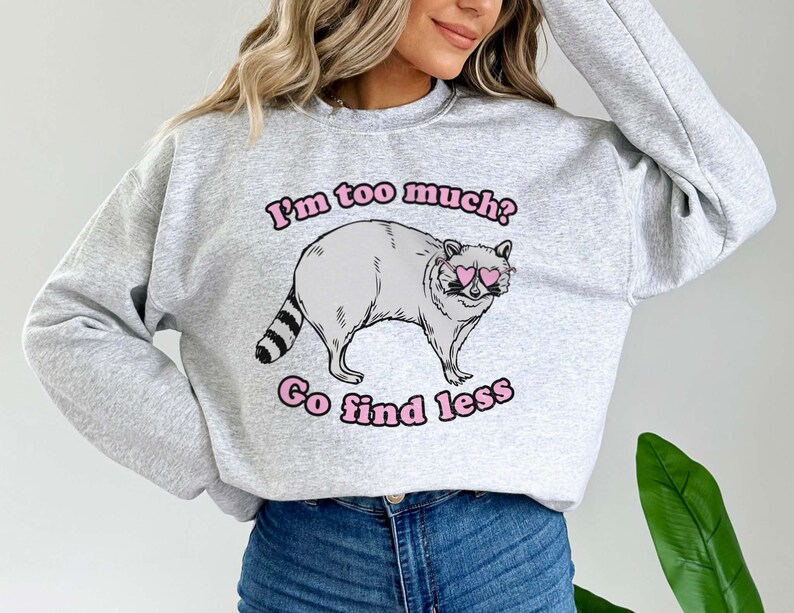 Pu&ograve; includere: Felpa grigia con un procione che indossa occhiali da sole a forma di cuore rosa e il testo "I'm too much? Go find less".