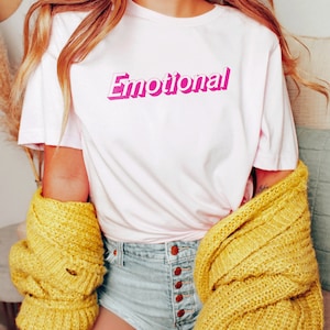 May include: A white t-shirt with the word "Emotional" printed in pink 3D lettering. The shirt is being worn by a person with long blonde hair and a yellow cardigan.