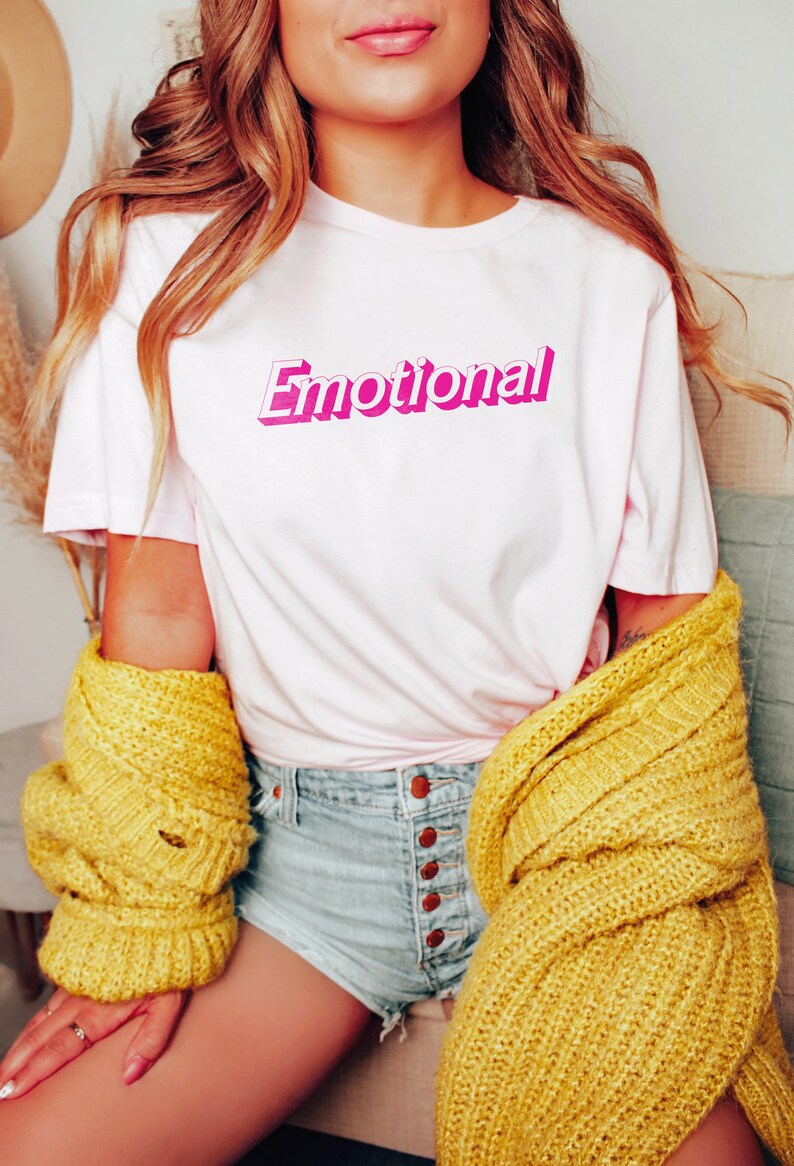 May include: A white t-shirt with the word "Emotional" printed in pink 3D lettering. The shirt is being worn by a person with long blonde hair and a yellow cardigan.