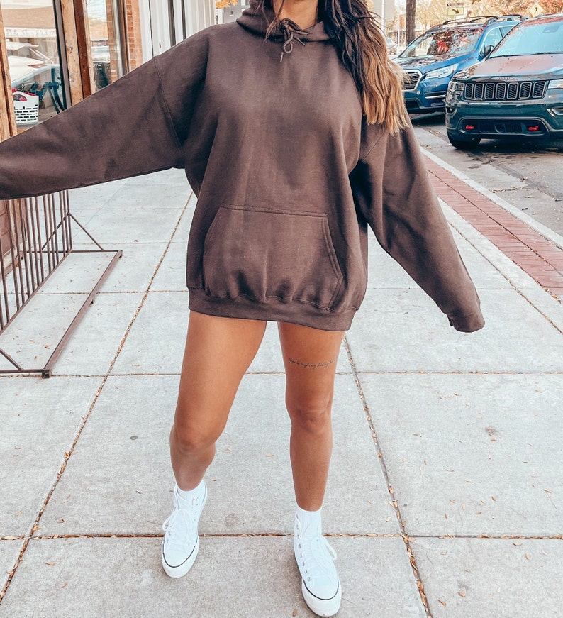 K&ouml;nnte beinhalten: Ein brauner Oversize-Hoodie mit einer Kapuze mit Kordelzug und einer K&auml;ngurutasche. Das Model tr&auml;gt wei&szlig;e High-Top-Sneaker.