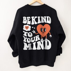 K&ouml;nnte beinhalten: Schwarzer Sweatshirt mit einem wei&szlig;en Grafikdruck, auf dem "Be Kind to Your Mind" steht. Die Grafik zeigt ein Cartoon-Herz mit Augen und einem Mund, das einen Finger zur Seite h&auml;lt.