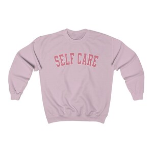 Peut inclure: Un sweat-shirt rose p&acirc;le avec le texte "SELF CARE" en lettres roses en arc de cercle.