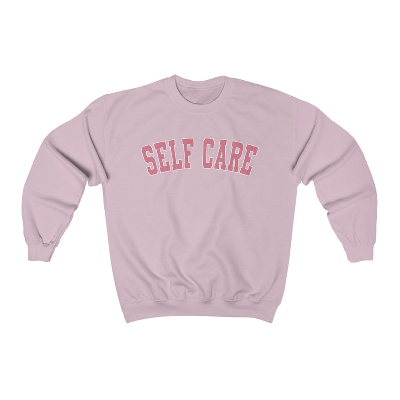 Peut inclure: Un sweat-shirt rose p&acirc;le avec le texte "SELF CARE" en lettres roses en arc de cercle.