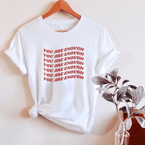 May include: White t-shirt with the phrase "YOU ARE ENOUGH" repeated in red lettering.