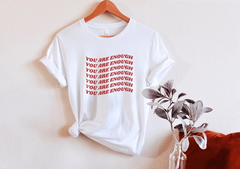 May include: White t-shirt with the phrase "YOU ARE ENOUGH" repeated in red lettering.