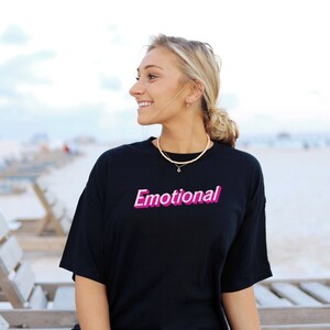 May include: A woman wearing a black t-shirt with the word "Emotional" printed in pink on the front. She is sitting on a wooden bench with a light blue sky in the background.