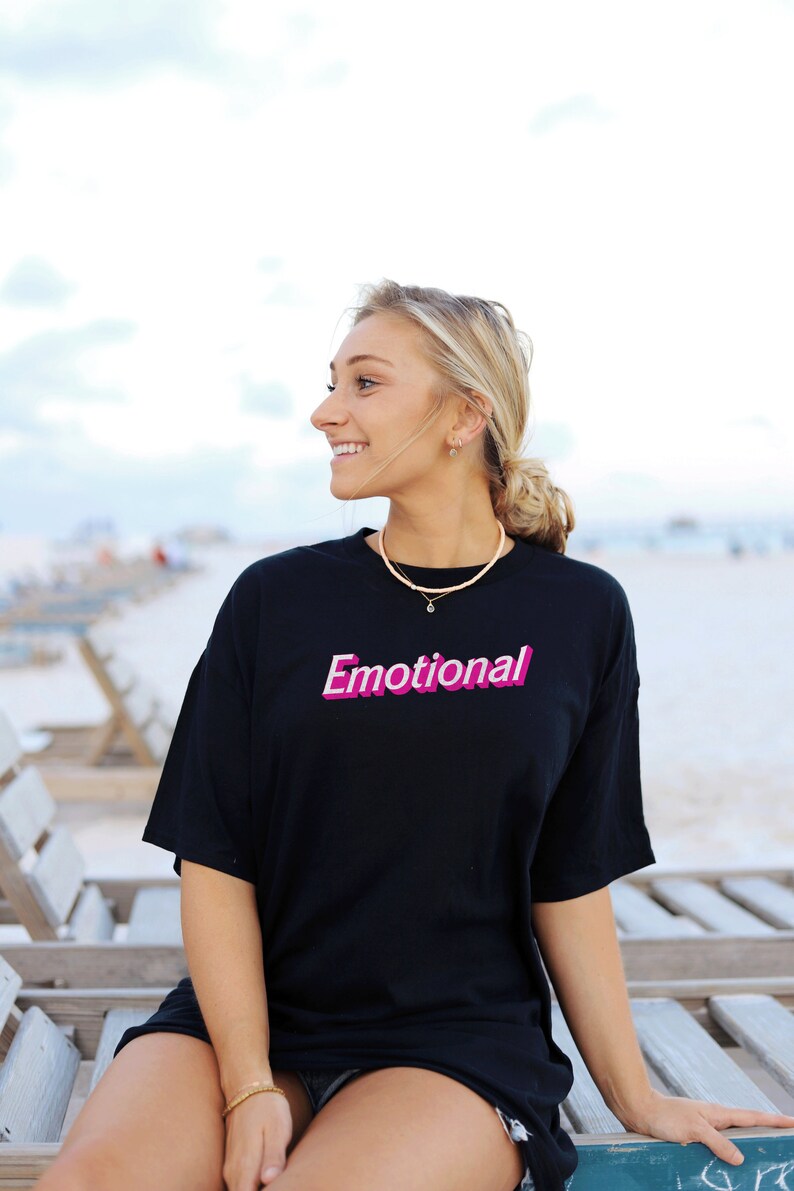 May include: A woman wearing a black t-shirt with the word "Emotional" printed in pink on the front. She is sitting on a wooden bench with a light blue sky in the background.