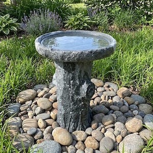 May include: A dark gray stone birdbath featuring a basin filled with water, supported by a pedestal designed to resemble a textured tree trunk. The birdbath is surrounded by a collection of smooth, rounded river stones of various sizes.