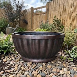 May include: A large, round, dark bronze-colored planter with a wide, flat rim and a vertically fluted exterior design. The bowl-shaped container features a smooth, rounded base and a deep, dark interior, presenting a classic, sturdy appearance.