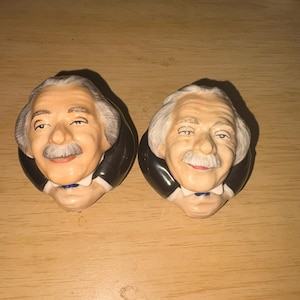 May include: Two ceramic salt and pepper shakers shaped like the head and shoulders of Albert Einstein. Each figure features gray hair, a mustache, a black jacket, a white shirt collar, and a small blue bowtie, rendered in a glossy finish.