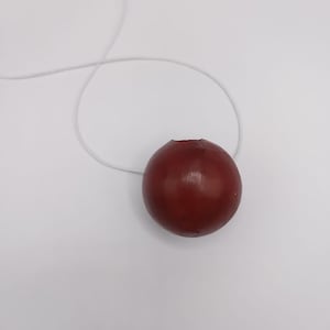 Puede incluir: Un objeto redondo, de color rojo oscuro, con una pequeña abertura en la parte superior, conectado a un hilo blanco y delgado. El objeto parece estar hecho de un material liso y se encuentra sobre un fondo blanco liso. El hilo está curvado y se extiende hacia arriba.