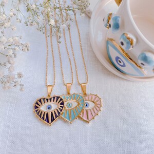 May include: Three gold necklaces with heart-shaped pendants featuring an evil eye design. The pendants are in shades of blue, pink, and purple.