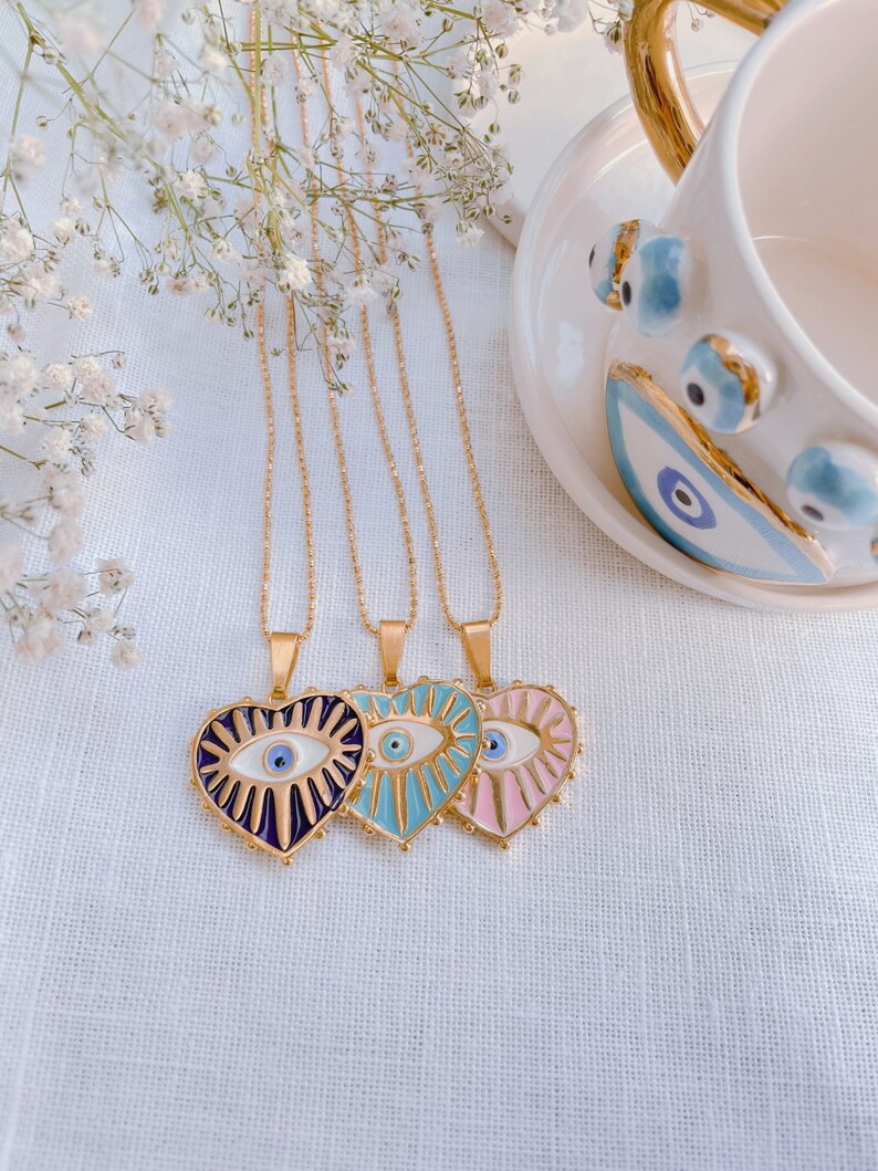 May include: Three gold necklaces with heart-shaped pendants featuring an evil eye design. The pendants are in shades of blue, pink, and purple.