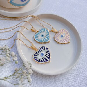 May include: Three gold necklaces with heart-shaped pendants featuring blue evil eyes. The pendants are decorated with white and blue enamel and have a gold rim. The necklaces are displayed on a white plate.