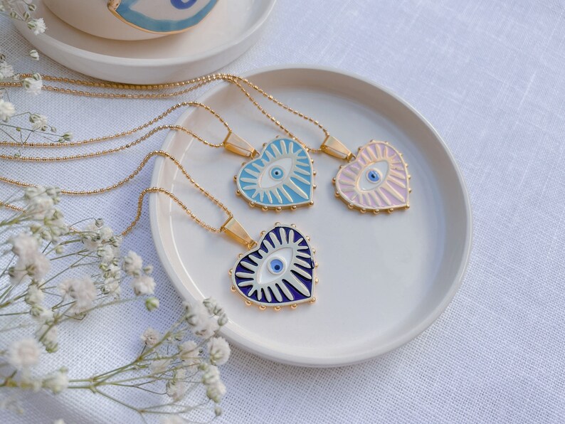 May include: Three gold necklaces with heart-shaped pendants featuring blue evil eyes. The pendants are decorated with white and blue enamel and have a gold rim. The necklaces are displayed on a white plate.