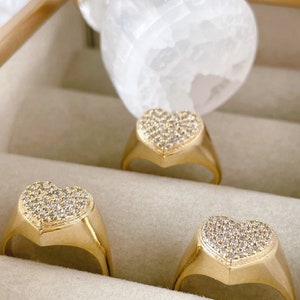 Könnte beinhalten: Drei goldene herzförmige Ringe mit klaren Zirkonia-Steinen. Die Ringe werden in einer samtenen Schmuckschatulle präsentiert.