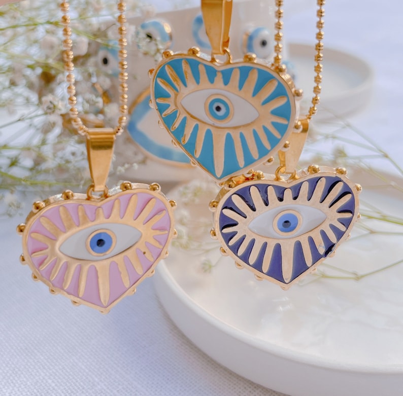 May include: Three gold necklaces with heart-shaped pendants featuring an evil eye design. The pendants are in shades of blue, pink, and purple.
