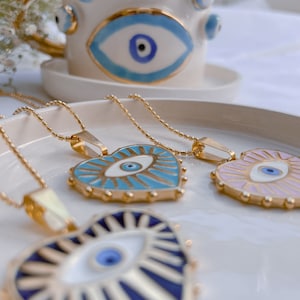 May include: Three gold necklaces with heart-shaped pendants featuring an evil eye design. The pendants are blue, white, and pink with gold accents.