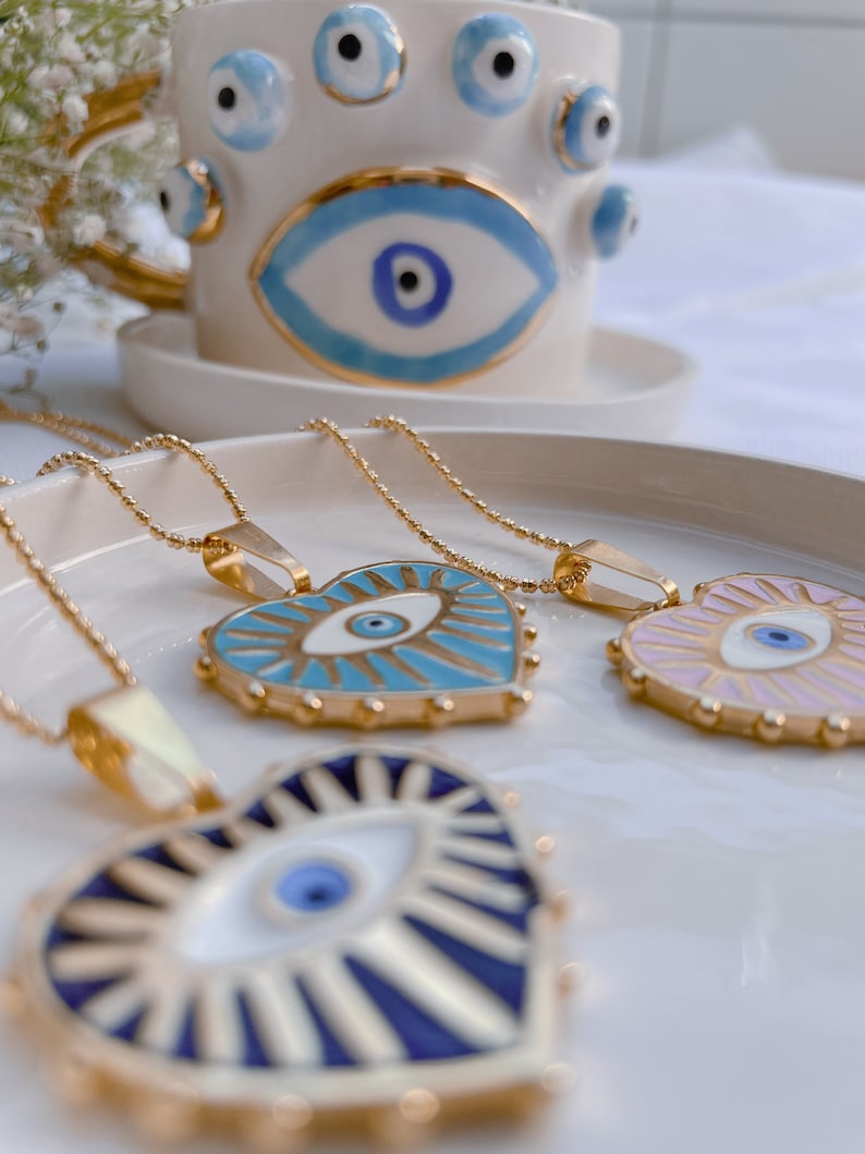 May include: Three gold necklaces with heart-shaped pendants featuring an evil eye design. The pendants are blue, white, and pink with gold accents.