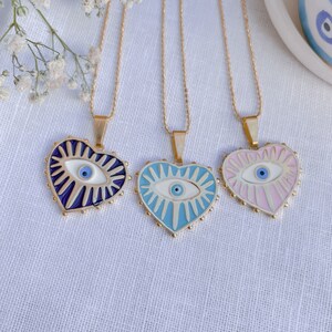 May include: Three gold chain necklaces with heart-shaped pendants featuring an evil eye design. The pendants are blue, light blue, and pink.