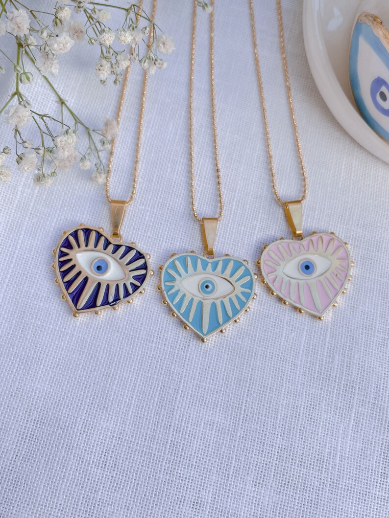 May include: Three gold chain necklaces with heart-shaped pendants featuring an evil eye design. The pendants are blue, light blue, and pink.