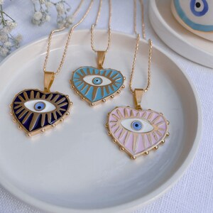 May include: Three gold necklaces with heart-shaped pendants featuring an evil eye design. The pendants are blue, pink, and black with gold accents.